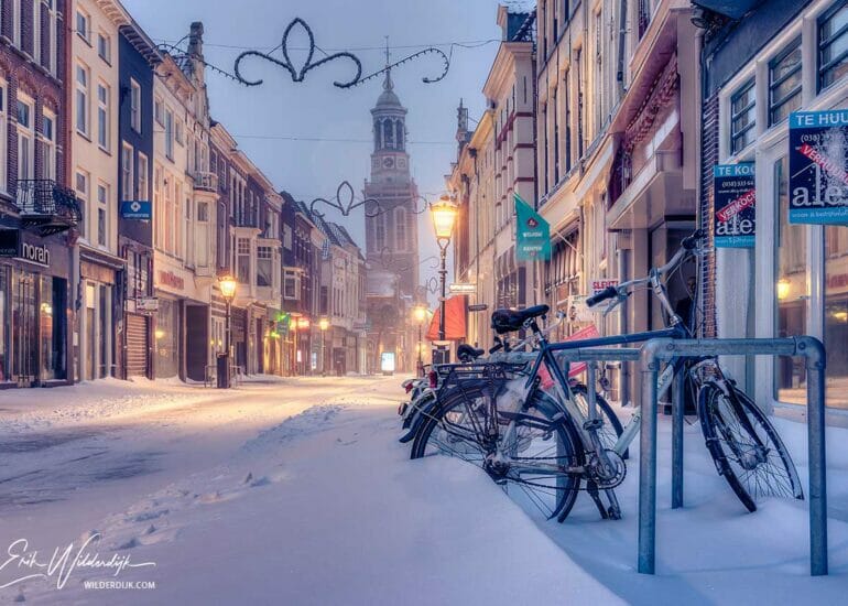 Een besneeuwde Oudestraat in Kampen met ingesneeuwde fietsen op de voorgrond en de Nieuwe Toren op de achtergrond